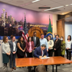 Mayor Bruce Harrell signs a bill while sitting at a table. A group of people stand behind him. The Washington and American flags are in the background. A picture of the Seattle skyline is in the background.