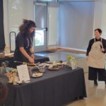 A chef cooks poblano peppers from an electric induction stove. The stove is on a tab le with a black tablecloth. White walls and a concrete floor are in the background. Another chef is holding a microphone and standing next to the table.