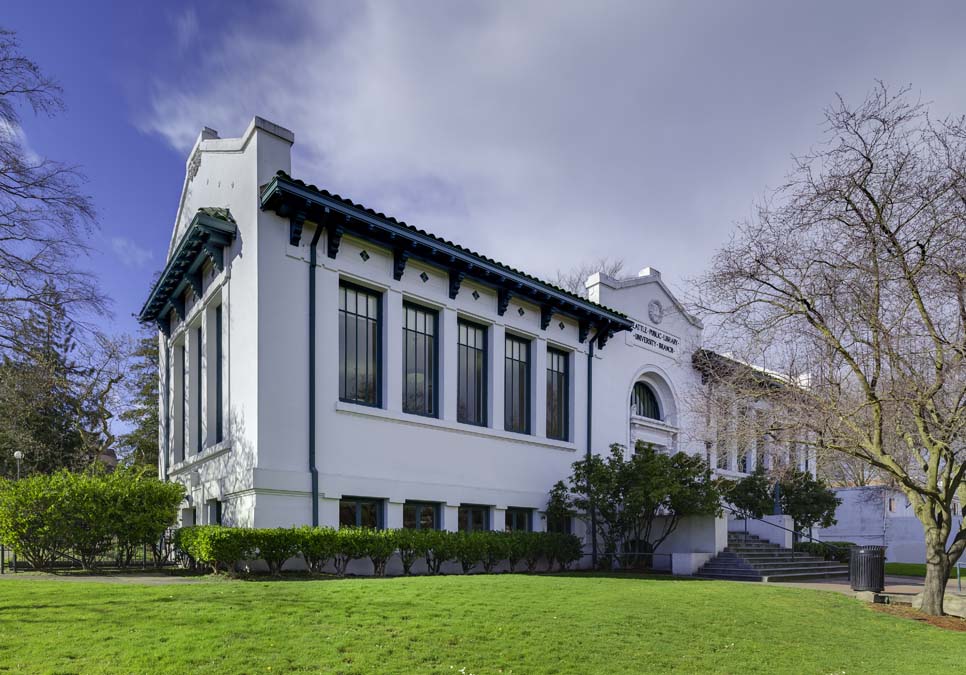 A white building with large rectangular windows and dark blue trim. In the foreground, a grass lawn with shrubs right next to the building. Stairs lead up to the main entrance of the building, above which a sign reads “Seattle Public Library, University Branch.”