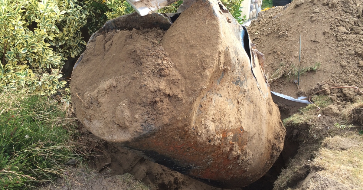 An old underground heating oil tank being lifted out of the ground by excavation equipment, covered in dirt