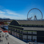 The Seattle Aquarium’s Pier 59 Building is in the foreground. It’s dark green with a light green trim, with a large sign reading “Seattle Aquarium” above large windows with white trim.
