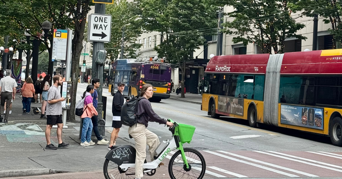 A person rides a bike past a bus stop as people wait for transit on a busy downtown Seattle street lined with trees and buildings