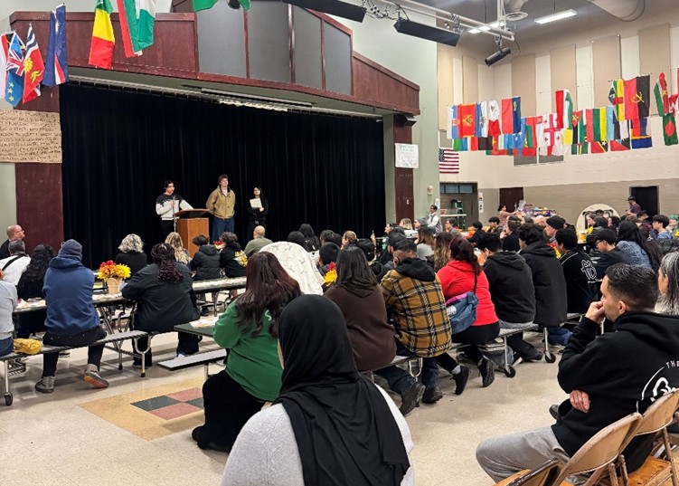 Concord Elementary gymnasium hosts DRCC’s annual Environmental Justice Forum. Youth Leaders address a filled gym from a stage with international flags hung overhead.