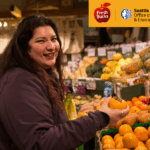 A woman is standing in front of bins filled with various fruits and vegetables, holding a persimmon.