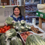 Store owner Anna Chen holding a bag of leafy green vegetables inside her store HC Grocery, surrounded by shelves stocked with produce, sauces, noodles, and more.