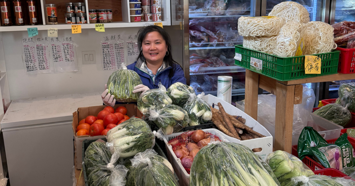 Store owner Anna Chen holding a bag of leafy green vegetables inside her store HC Grocery, surrounded by shelves stocked with produce, sauces, noodles, and more.