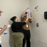 Students stand in the shower room, one holds a plastic bag with lines on it used to measure the water efficiency of the shower head, while two other students help read and record the measurements.