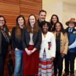 Group of professionals standing indoors against a wooden wall, dressed in business casual attire, posing for a photo.