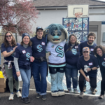 A group of City of Seattle staff wearing blue Kraken shirts, smiling next to the Seattle Kraken’s troll mascot on an outdoor basketball court at the South Park Community Center and Playfield.