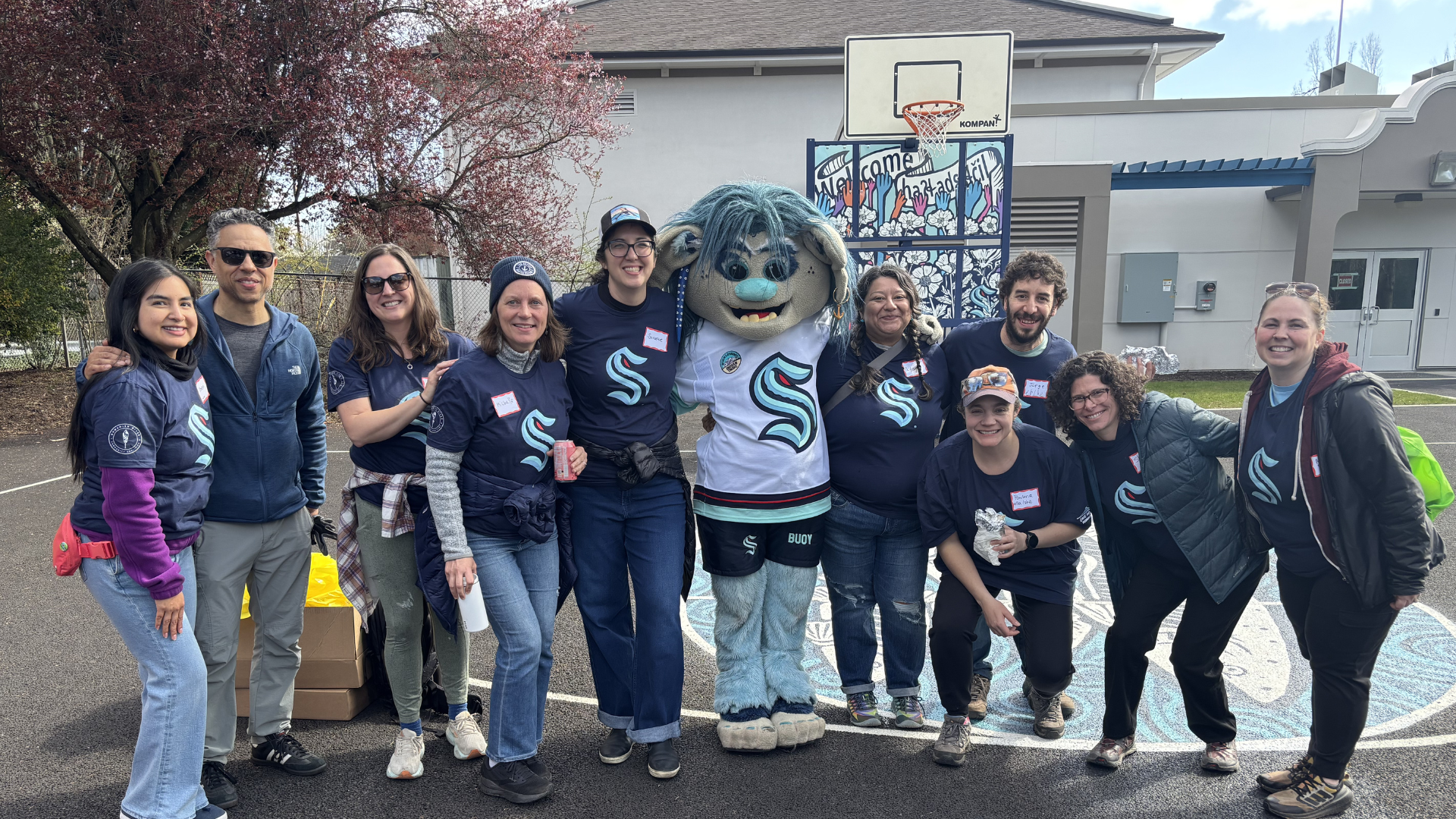 A group of City of Seattle staff wearing blue Kraken shirts, smiling next to the Seattle Kraken’s troll mascot on an outdoor basketball court at the South Park Community Center and Playfield.