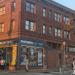 People walking on the street in front of a red brick building on a sunny day.