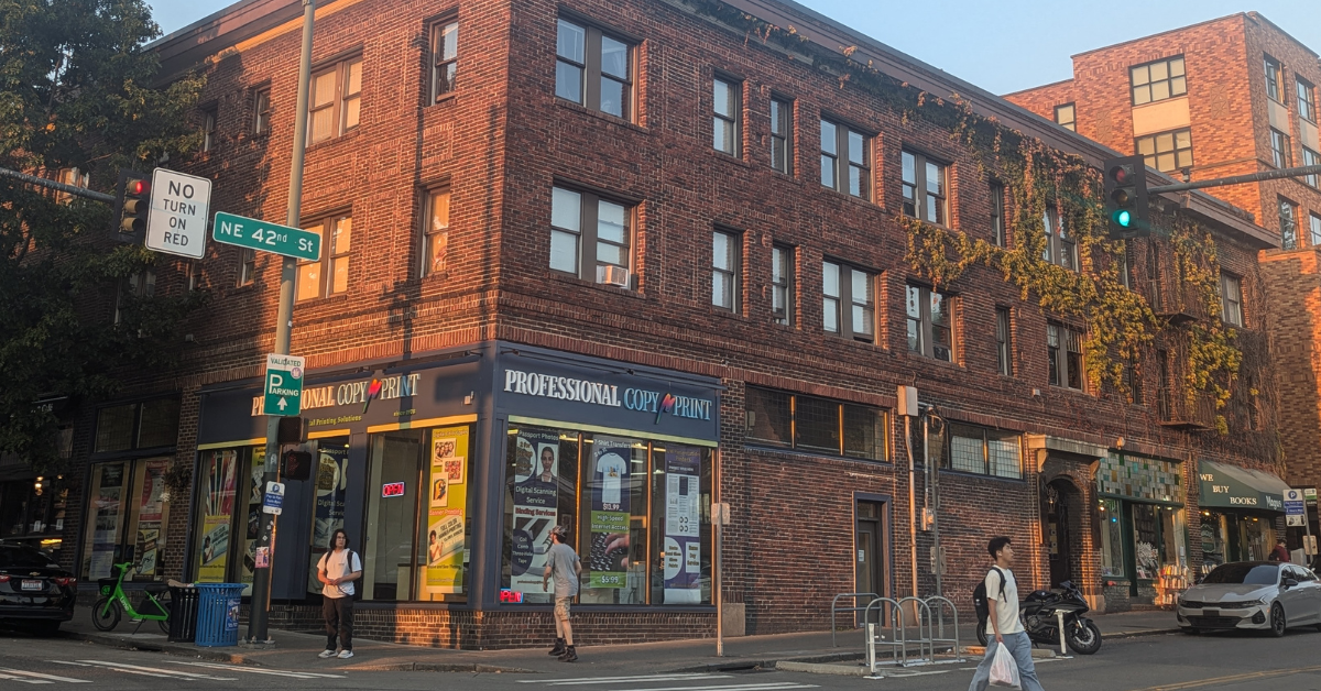 People walking on the street in front of a red brick building on a sunny day.