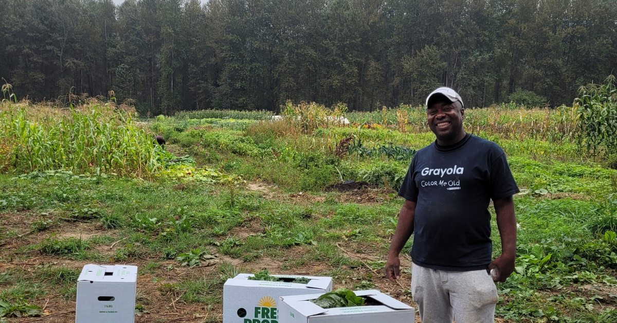A farmer smiles near three fresh produce boxes in front of green grass and trees.