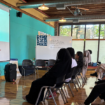 A presenter stands at the front of the room in front of two rows of participants sitting in chairs at the Youth Community Assembly