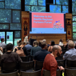 An audience seated in a wood-paneled room facing a large screen reading ‘Welcome to the Seattle Regional Partners Forum.’