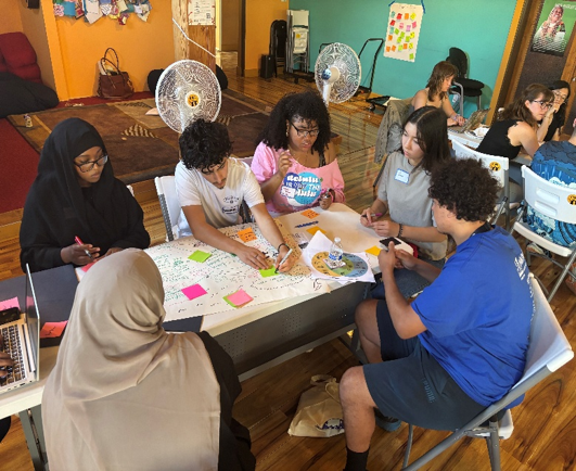 A group of young adults sit around a table during a group activity, writing on sticky notes and a poster.