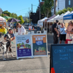 Shoppers interacting at a farmers market, surrounded by vibrant stalls offering a variety of local products and fresh foods, signage advertising Fresh Bucks and SNAP-EBT.