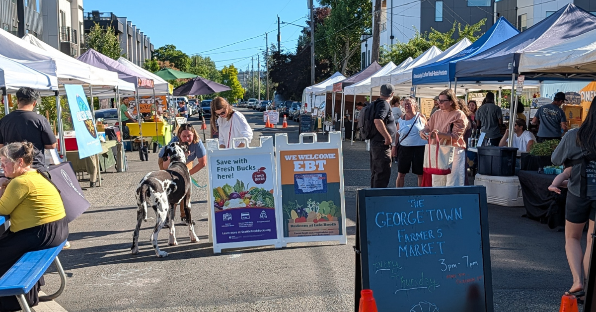 Shoppers interacting at a farmers market, surrounded by vibrant stalls offering a variety of local products and fresh foods, signage advertising Fresh Bucks and SNAP-EBT.