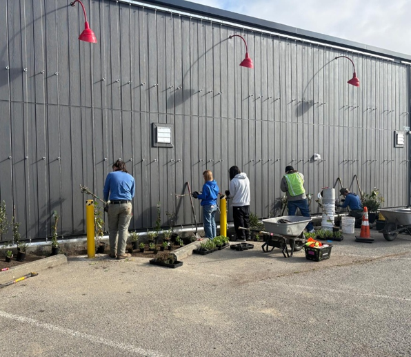A group of people standing in front of a large dark-colored building, gathered outdoors on a sunny day with tools and supplies visible nearby.