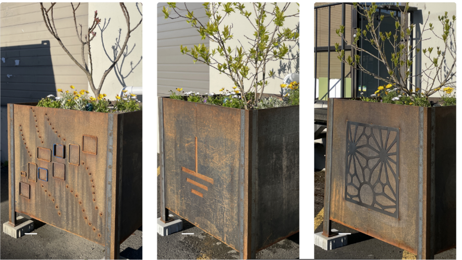 Three metal planters hold young trees and small plants, each decorated with a different cut‑out design, placed beside a building.